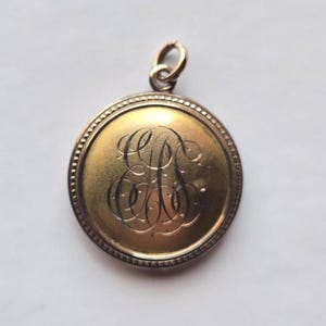 Könnte beinhalten: Ein goldfarbener runder Anhänger mit einem eingravierten Monogramm. Der Anhänger hat einen Perlenrand und eine kleine Schlaufe oben für eine Kette. Das Monogramm ist in einem eleganten Schriftstil gehalten.