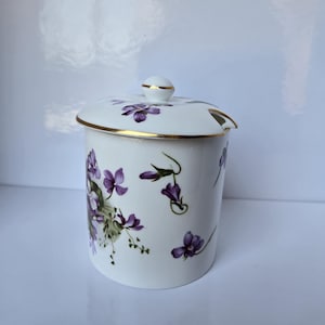 Puede incluir: Azucarero de cerámica blanca con tapa, decorado con diseños florales morados y verdes. La tapa tiene un borde dorado y un pequeño pomo.