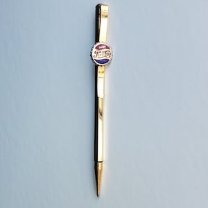 May include: A gold-colored pen with a Pepsi Cola bottle cap design on the top. The cap is red, white, and blue with the words "Drink Pepsi Cola" on it.