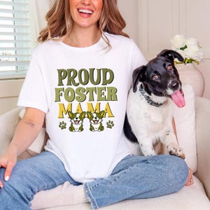 Op de afbeelding: Een wit t-shirt met korte mouwen voorzien van de tekst PROUD FOSTER MAMA in olijfgroene en gele letters, vergezeld door een afbeelding van twee kleine bruin-witte honden en vier pootafdrukken in het midden van het kledingstuk.