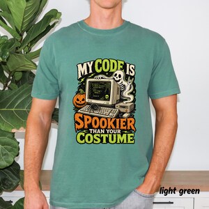 May include: A light green t-shirt featuring a graphic of a computer, a skeleton, a pumpkin, and a spider. The text reads "MY CODE IS SPOOKIER THAN YOUR COSTUME". The design includes a vintage monitor displaying code, a keyboard, and a mouse.