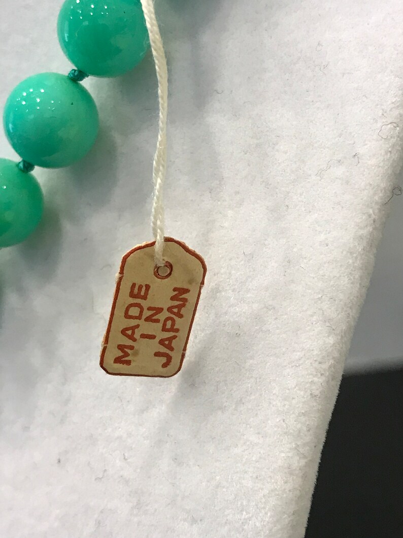 May include: Close-up of a turquoise beaded necklace with a tag that reads "MADE IN JAPAN". The necklace features large, round beads. The tag is rectangular with rounded corners and a string attached.