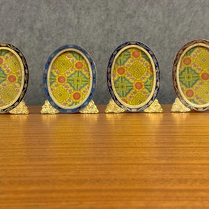 May include: Four oval-shaped picture frames with gold-colored ornate metal stands. Each frame features a dark-colored border and contains a yellow insert with a repeating pattern of pink and green floral motifs and geometric lines.