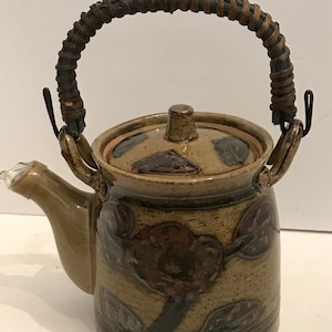 May include: A ceramic teapot with a brown and tan floral design. The teapot has a woven handle and a spout with a clear tip. The lid has a small knob. The teapot is a decorative item for the kitchen.