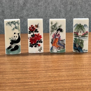 May include: Four rectangular decorative tiles with Asian-inspired artwork. The tiles feature images of a panda, red flowers, a woman in traditional dress, and a landscape with mountains and trees. The tiles are cream-colored and have a glossy finish.