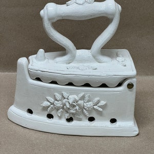 May include: A white decorative iron with a handle and floral embellishments. The iron features a detailed rose design on the front and is made of a solid, possibly ceramic, material. The handle has a curved design.