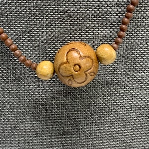 May include: Wooden bead necklace featuring a large, carved flower pendant. The pendant is flanked by two smaller, round beads. The necklace is made of small, brown wooden beads. The background is a textured gray fabric.