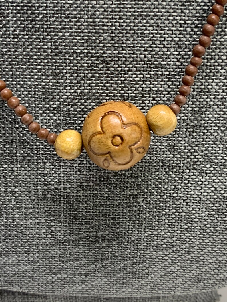 May include: Wooden bead necklace featuring a large, carved flower pendant. The pendant is flanked by two smaller, round beads. The necklace is made of small, brown wooden beads. The background is a textured gray fabric.