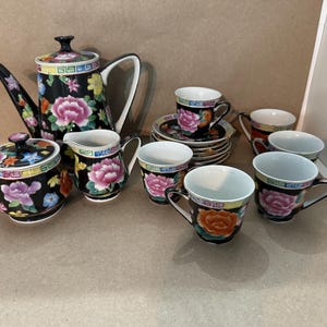 May include: A black porcelain tea set with floral designs. The set includes a teapot, sugar bowl, creamer, and several teacups and saucers. The floral pattern features pink, orange, and yellow flowers.
