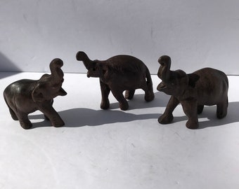 May include: Three small, dark brown elephant figurines. Each elephant has its trunk raised and is standing on four legs. The figurines are made of a solid material and are displayed on a white surface. The elephants are a decorative item.