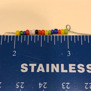 May include: A strand of colorful beads, including yellow, green, red, white, black, and blue, strung on a thin wire. The beads are approximately 2.5 inches long, as measured by a blue ruler.
