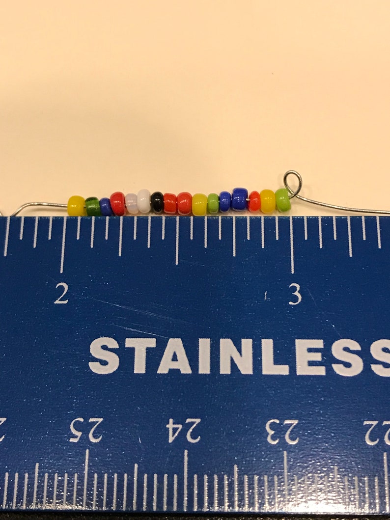May include: A strand of colorful beads, including yellow, green, red, white, black, and blue, strung on a thin wire. The beads are approximately 2.5 inches long, as measured by a blue ruler.