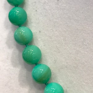 May include: Close-up of a necklace featuring large, round, light-green beads strung together with matching green thread. The beads have a glossy finish and are evenly spaced, creating a simple yet elegant design. The background is a light, neutral color.