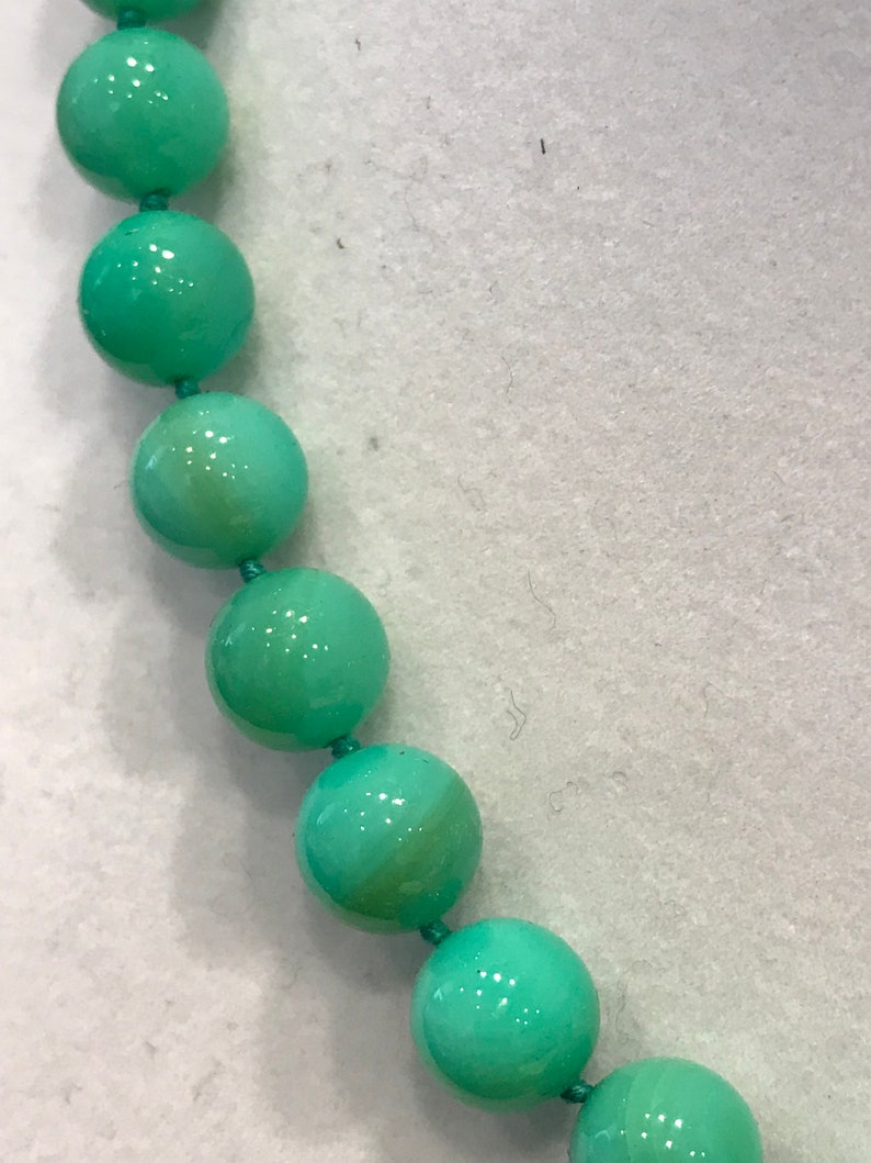 May include: Close-up of a necklace featuring large, round, light-green beads strung together with matching green thread. The beads have a glossy finish and are evenly spaced, creating a simple yet elegant design. The background is a light, neutral color.