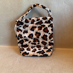 May include: A small, leopard print bag with a handle. The bag is a soft, plush material with a repeating pattern of brown spots on a cream background. The handle is the same leopard print. The bag has a drawstring closure.