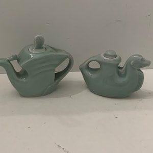 May include: Two light green ceramic teapots. One is a traditional teapot shape, and the other is shaped like a duck. Both have a glossy finish and are set against a white background.