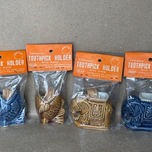 May include: Four ceramic toothpick holders in clear plastic packaging. The holders are shaped like an owl, elephant, and other figures, in blue, brown, and tan colors. Each holder has a small opening for toothpicks and is labeled "Ceramic Toothpick Holder".