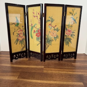 May include: A four-panel folding screen featuring dark brown wooden frames with intricate cut-out designs at the base. Each panel displays a light yellow background with painted illustrations of colorful birds perched on flowering branches.