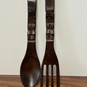 May include: A pair of dark brown wooden serving utensils consisting of a large spoon and a four-tined fork. Each handle features a carved face design and a small circular hole at the top for hanging. The wood has a smooth, polished finish.