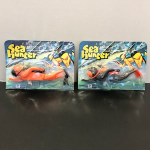 May include: Two vintage Sea Hunter wind-up swimming action toys in their original packaging. One toy is orange, the other is black. The packaging features the words "Sea Hunter" in yellow and blue, with an illustration of a diver.