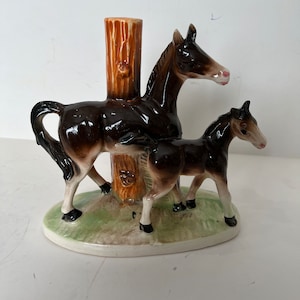 May include: A ceramic horse figurine featuring a mare and foal standing on a green and white oval base. The horses are brown with black manes and tails, and pink noses. A brown tree trunk is between the horses.