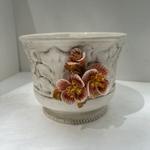 May include: A decorative, off-white ceramic planter with embossed leaf patterns and a raised base. The planter features three-dimensional, reddish-brown and yellow flower accents. The rim has a slightly flared shape.