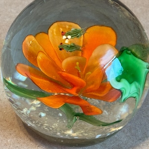 May include: A clear glass paperweight with an orange flower and two small green frogs inside. The flower has orange petals and green stems and leaves. The frogs have red eyes.