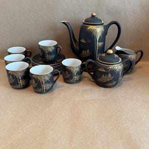 May include: A black and gold tea set featuring a teapot, sugar bowl, creamer, saucers, and cups. The set has a bamboo and landscape design in gold. The cups and saucers are stacked on a brown surface.