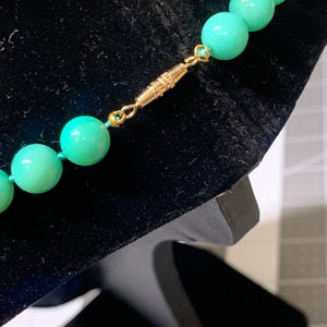 May include: Close-up of a turquoise beaded necklace with a gold-colored clasp. The beads are round and evenly spaced, strung on a light blue cord. The clasp is a simple, rectangular design. The necklace is displayed on a black velvet surface.