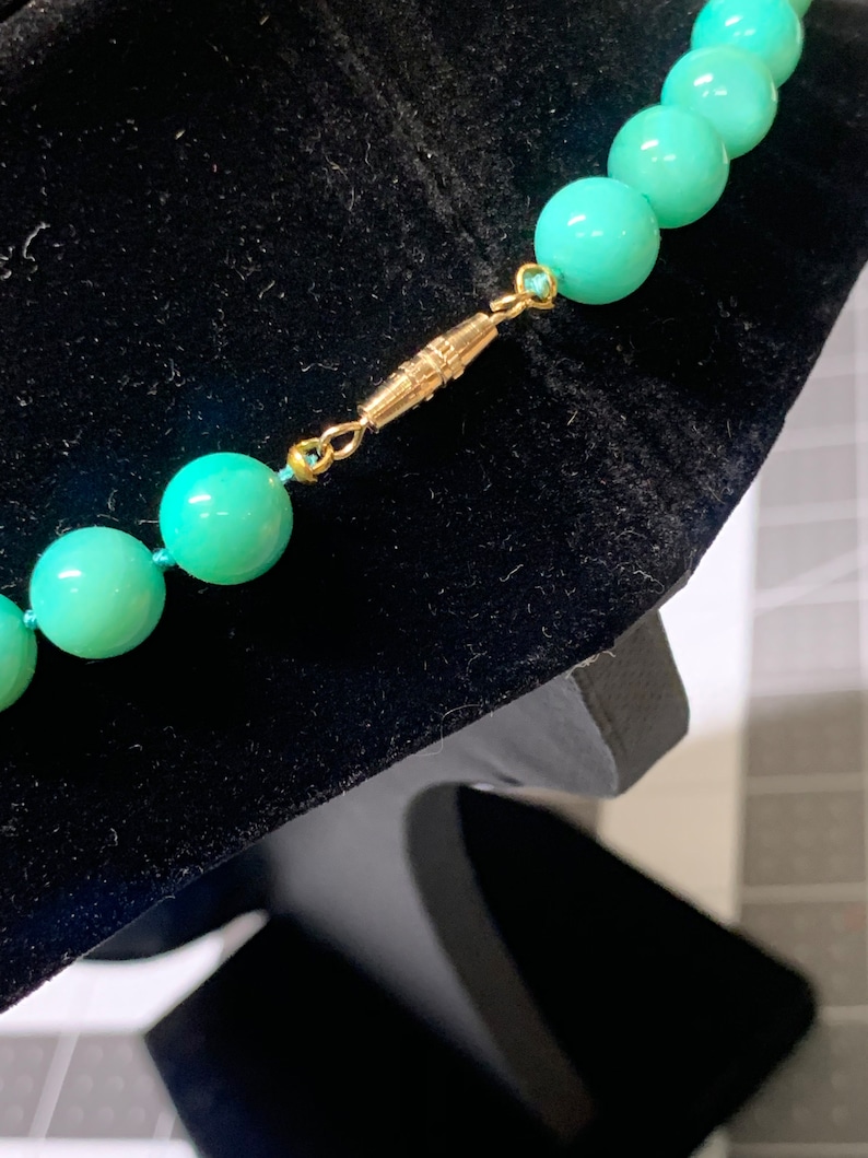May include: Close-up of a turquoise beaded necklace with a gold-colored clasp. The beads are round and evenly spaced, strung on a light blue cord. The clasp is a simple, rectangular design. The necklace is displayed on a black velvet surface.