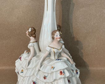 May include: A white porcelain figurine featuring three women in tiered dresses, adorned with floral accents. The central element is a tall, slender vase with gold trim. The base is round and the overall design is elegant and detailed.