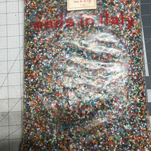 May include: A clear plastic bag filled with small, colorful beads. The beads are a mix of colors including red, orange, yellow, blue, and white. The bag has a label that reads "Col. N. Multicolor" and "Size N. 2/0". The text "made in italy" is visible.
