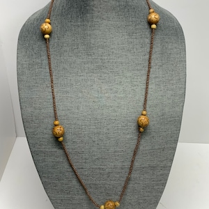 May include: A long necklace with a brown cord and alternating small beads and larger, light-colored wooden beads. The wooden beads have a carved floral design. The necklace is displayed on a gray mannequin.