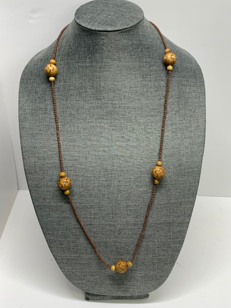 May include: A long necklace with a brown cord and alternating small beads and larger, light-colored wooden beads. The wooden beads have a carved floral design. The necklace is displayed on a gray mannequin.