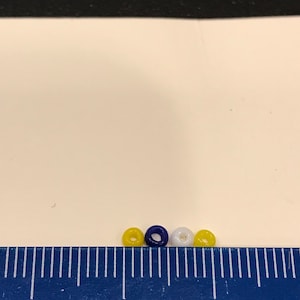 May include: Four small, round beads in yellow, navy blue, and white are arranged in a row. A blue ruler with white markings is in the foreground, with the beads positioned between the 2 and 3-inch marks. The background is white.