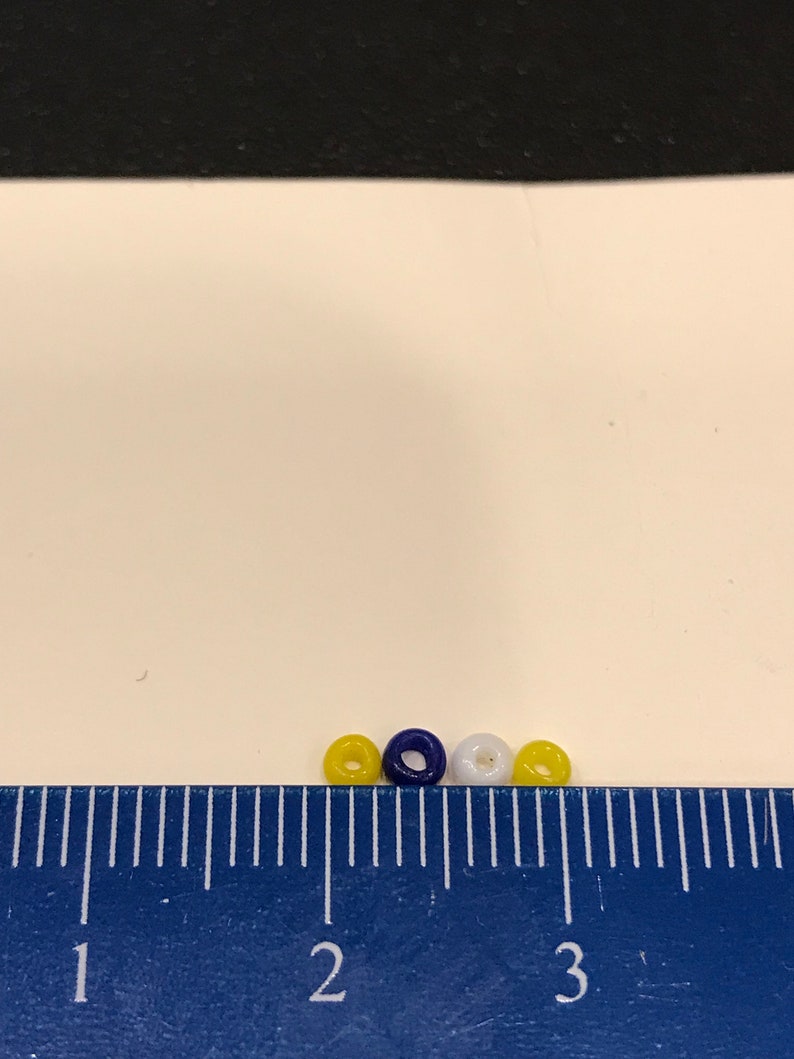 May include: Four small, round beads in yellow, navy blue, and white are arranged in a row. A blue ruler with white markings is in the foreground, with the beads positioned between the 2 and 3-inch marks. The background is white.