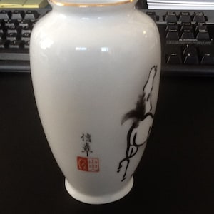 May include: White ceramic vase with a gold rim and a black and white horse illustration. The vase has Japanese characters and a red seal. The vase is a decorative item.