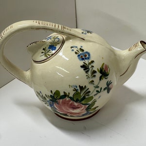 May include: Cream-colored ceramic teapot with a curved handle and spout. The teapot is decorated with hand-painted floral designs in shades of pink, blue, and green. The handle has brown stripes. The teapot sits on a white surface.