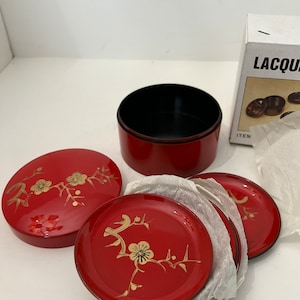 May include: A set of red lacquered serving dishes with gold floral designs. The set includes a round container, a lid, and several small plates. The plates and lid feature a gold floral pattern. The interior of the container is black.