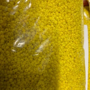 May include: A clear plastic bag filled with small, yellow seed beads. The beads are uniform in size and shape, with a slightly rounded appearance. The bag is partially visible, showing the beads' texture and color.