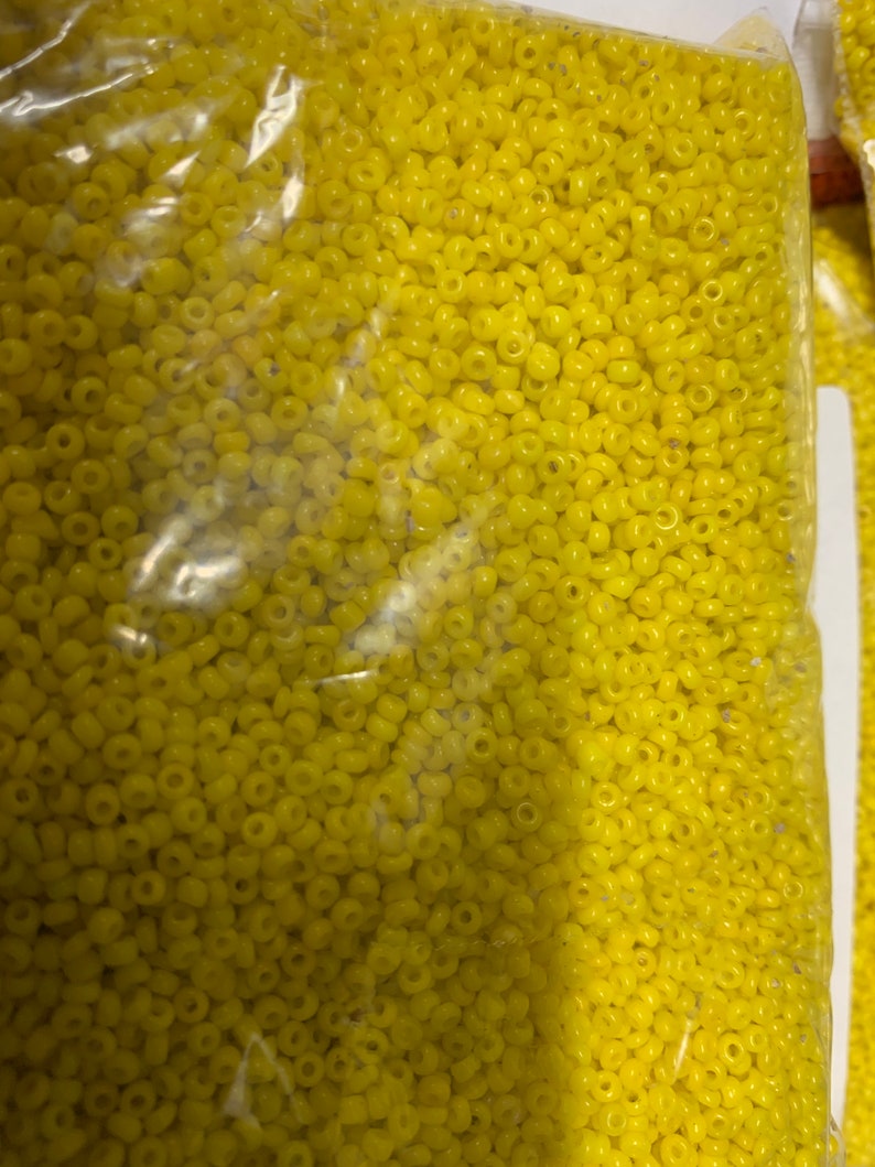 May include: A clear plastic bag filled with small, yellow seed beads. The beads are uniform in size and shape, with a slightly rounded appearance. The bag is partially visible, showing the beads' texture and color.