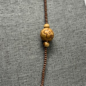 May include: A long necklace featuring small brown beads and a large wooden bead with a carved flower design. The necklace is displayed against a gray textured background.