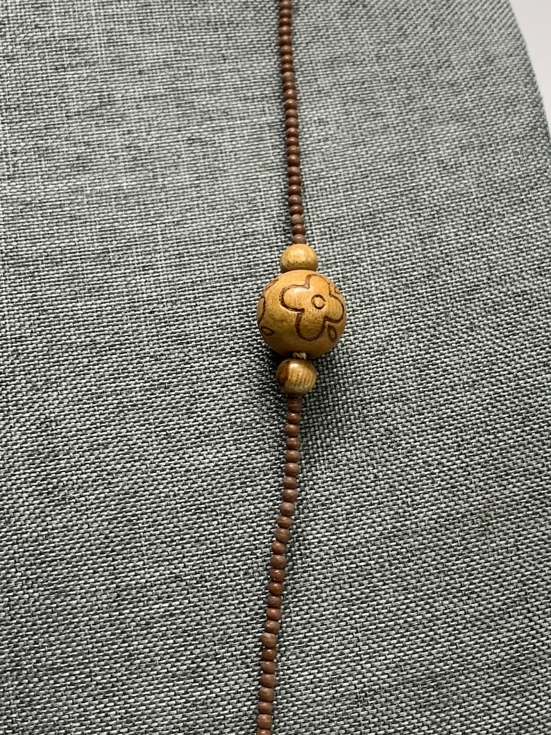 May include: A long necklace featuring small brown beads and a large wooden bead with a carved flower design. The necklace is displayed against a gray textured background.