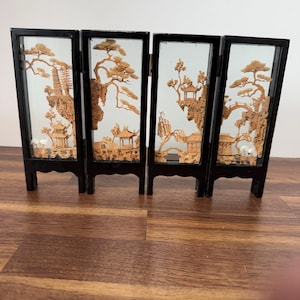 May include: A four-panel folding screen featuring intricate cork carvings of landscapes with pagodas, trees, and rock formations encased in black frames. Each panel displays a detailed scene with light brown natural cork textures against a clear background.