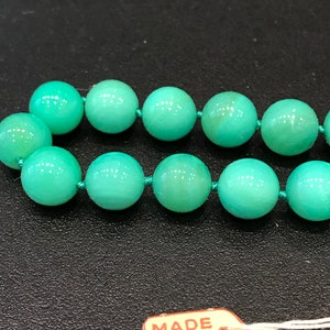 May include: Close-up of a necklace featuring round, light green beads strung on a matching green cord. The beads have a smooth, polished appearance. A small tag that reads "MADE IN JAPAN" is attached to the necklace.
