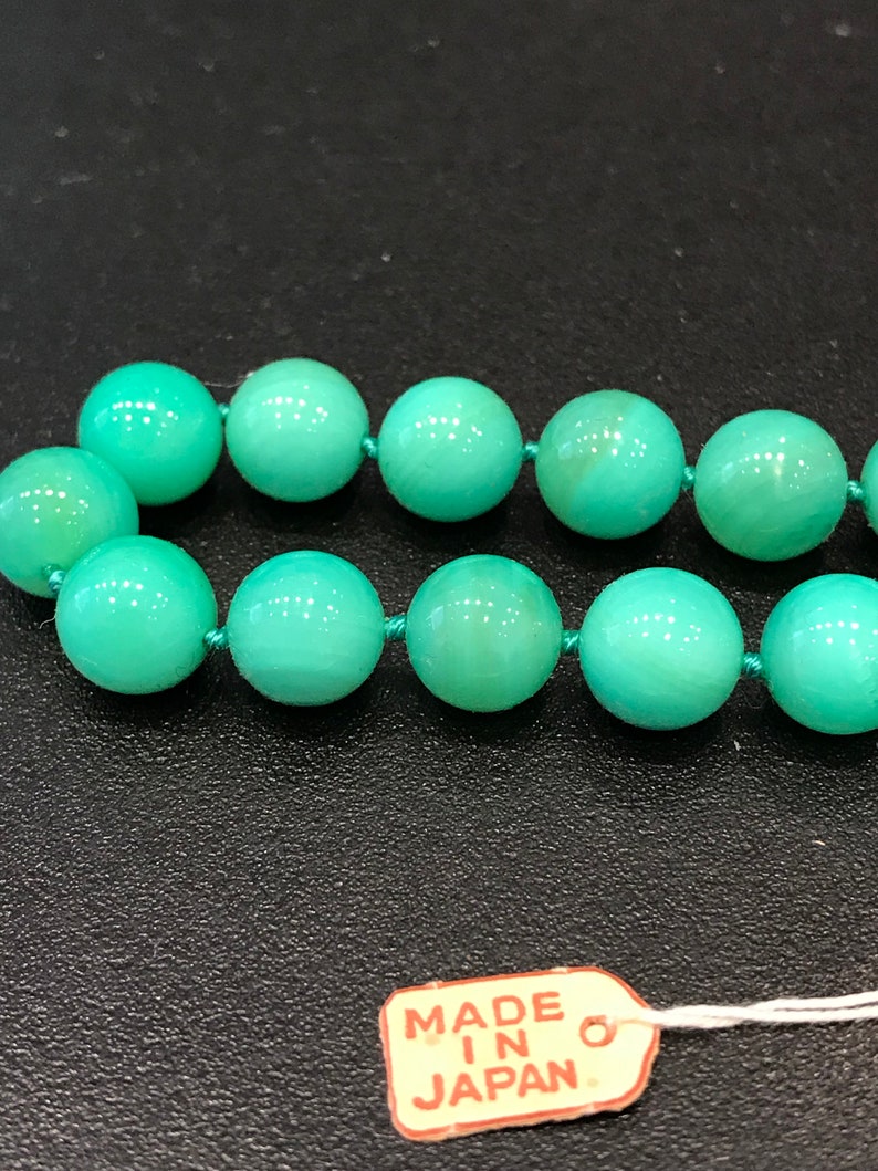 May include: Close-up of a necklace featuring round, light green beads strung on a matching green cord. The beads have a smooth, polished appearance. A small tag that reads "MADE IN JAPAN" is attached to the necklace.