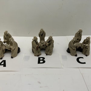 May include: Three sets of small, beige-colored figurines depicting elderly men in traditional robes. Each set shows the men gathered around a table or engaged in activities. The figurines are on a white surface.