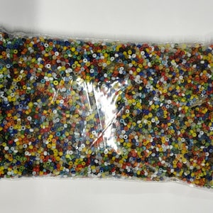 May include: A clear plastic bag filled with a large quantity of small, colorful beads. The beads are a mix of colors including red, yellow, blue, green, and white. The beads are small, round, and appear to be made of glass or plastic.