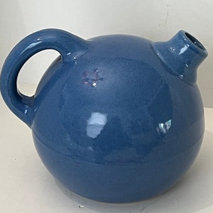 May include: A blue ceramic pitcher with a round body, a curved handle, and a spout. The pitcher has a glossy finish and a simple, elegant design. The handle is large enough to easily grip. The spout is small and angled.