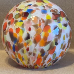 May include: A decorative glass sphere with a white base and colorful speckles of orange, yellow, blue, and brown. The speckled pattern creates a unique and artistic design. This sphere is a decorative accent piece.